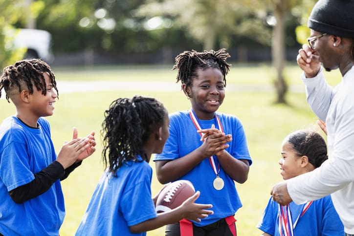 An 8 year old African-American girl smiling, standing with her teammates, with a medal hanging around her neck. The coach, her father, is giving out awards to the winning flag football team.